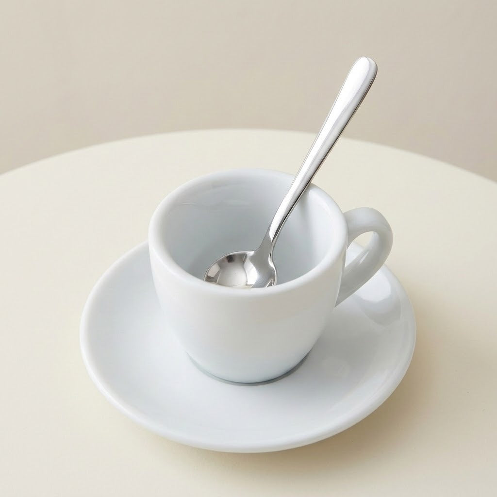 A white ceramic espresso cup with a matching saucer and a silver metal spoon, set on a light beige surface against a neutral background.