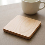A square light wood coaster with rounded corners sits on a neutral gray surface, with a white ceramic mug blurred in the background.