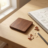 A square wooden block with rounded corners and a dark brown wood grain finish, resting on a light wood desk next to a spiral notebook, pencil, and wood shavings.