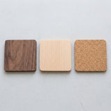 Three square coasters with rounded corners arranged horizontally on a light gray surface. The left coaster is dark brown wood with visible grain, the center coaster is light natural wood, and the right coaster is cork in a tan color.