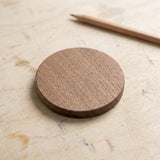 A round wooden coaster with a natural wood grain finish, placed on a light-colored wooden tabletop next to a wooden pencil.