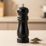 A black pepper mill grinder with a silver metal top knob, standing on a light beige countertop with a small pile of ground pepper in front of it. The background is a softly blurred kitchen setting.