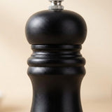 A close-up shot of a black pepper mill grinder, showing its textured matte black body and a silver metal top button against a soft beige background.