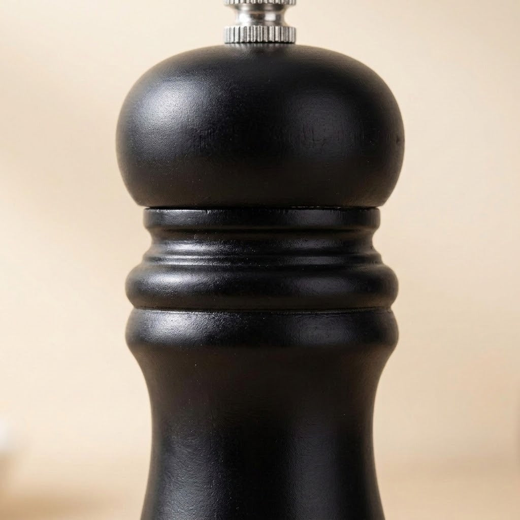 A close-up shot of a black pepper mill grinder, showing its textured matte black body and a silver metal top button against a soft beige background.