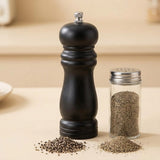 A black pepper mill grinder with a silver metal top knob stands next to a clear glass shaker bottle filled with coarse black pepper, with small piles of pepper scattered in front, all on a warm beige surface.