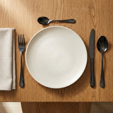 Glossy black cutlery surrounds a white plate on a warm wooden table beside a folded beige napkin.