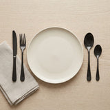 Black cutlery flanks a cream plate on a light wooden table, with a folded beige napkin beneath the knife and fork.