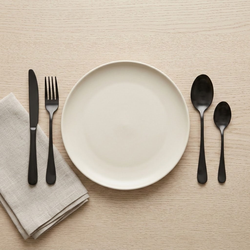 Black cutlery flanks a cream plate on a light wooden table, with a folded beige napkin beneath the knife and fork.