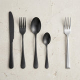 A matte black knife, fork, tablespoon and teaspoon lie beside a silver fork on a textured cream surface.