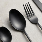 A close-up shows a matte black tablespoon and fork with a partially visible teaspoon and knife on a pale textured surface.