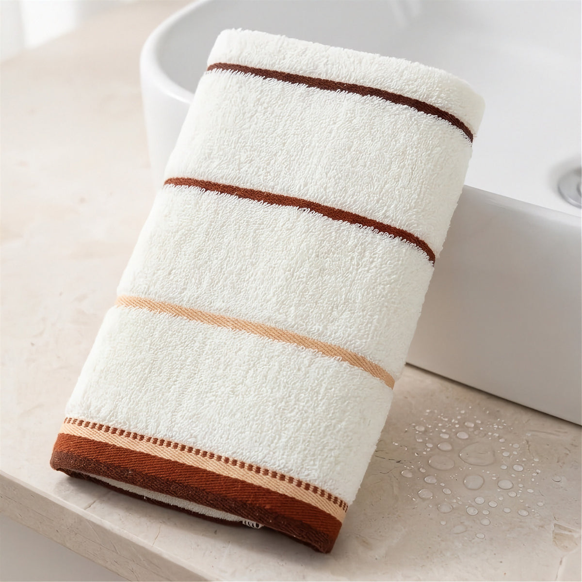 A folded cream terry towel with brown and beige stripes leans against a white sink on a countertop dotted with water.