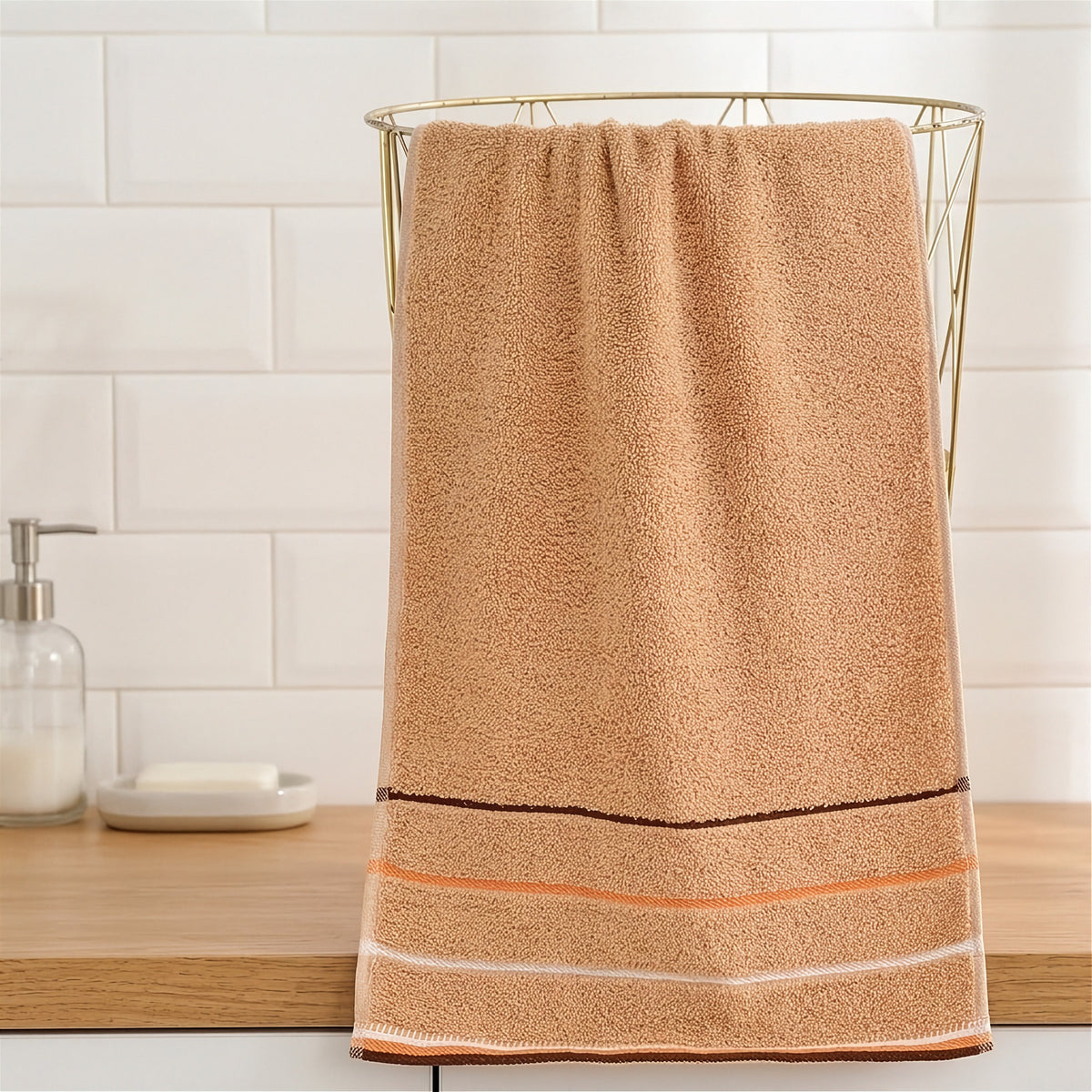 A tan terry towel with brown, peach, and white stripes hangs over a gold wire basket on a wooden bathroom counter.