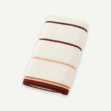 A folded white terry towel with thin brown and beige stripes and a brown border rests on an off-white background.