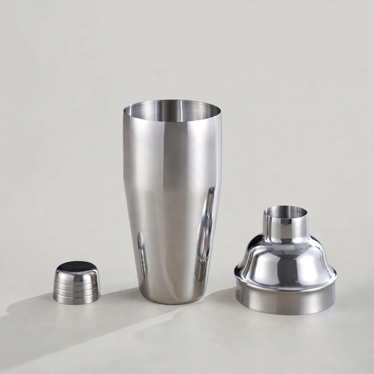 A stainless steel cocktail shaker set with a tall shaker body, a separate dome-shaped strainer, and a small black-and-silver cap, photographed on a clean light gray studio background.