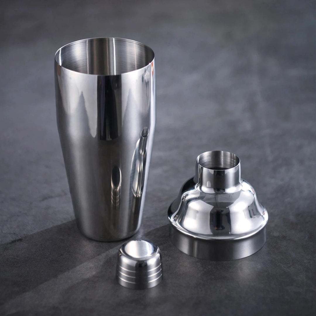 A polished silver cocktail shaker stands disassembled into a cup, domed lid, and small cap on a dark gray surface.