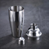A polished silver cocktail shaker stands disassembled into a cup, domed lid, and small cap on a dark gray surface.