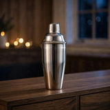 A polished silver cocktail shaker stands on a wooden counter with blurred warm lights and a dark window behind it.
