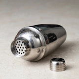 A silver cocktail shaker lies on a pale speckled countertop with its perforated strainer exposed and its cap beside it.