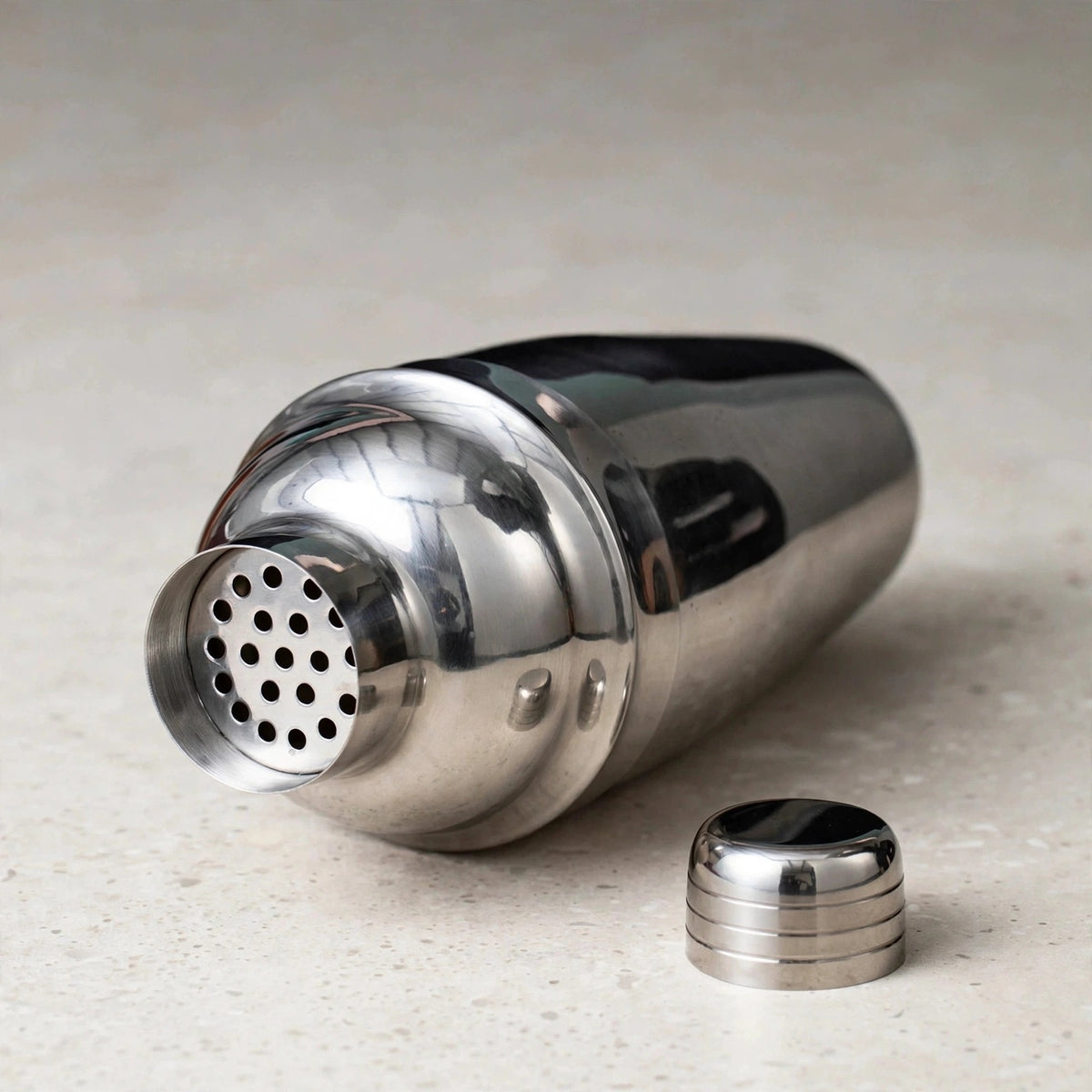 A silver cocktail shaker lies on a pale speckled countertop with its perforated strainer exposed and its cap beside it.