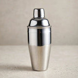 A polished silver cocktail shaker with a domed lid and grooved cap stands on a pale speckled countertop against a beige background.