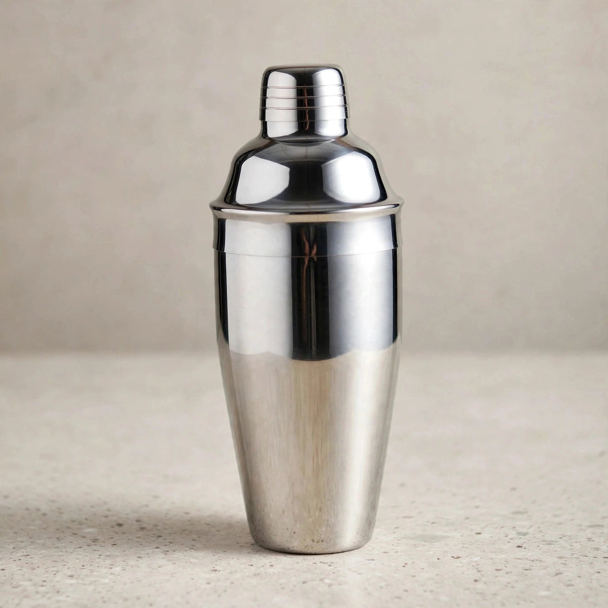 A polished silver cocktail shaker with a domed lid and grooved cap stands on a pale speckled countertop against a beige background.