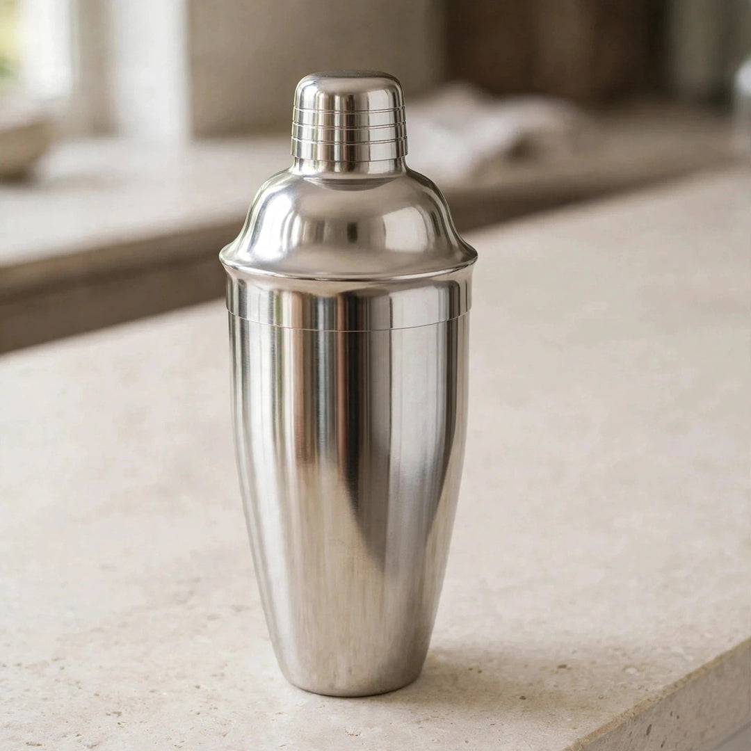 A polished stainless steel cocktail shaker standing on a light stone countertop with a softly blurred kitchen background.