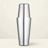 Polished stainless steel cocktail shaker with two tins fitted together stands upright on a cream surface against a pale cream background.