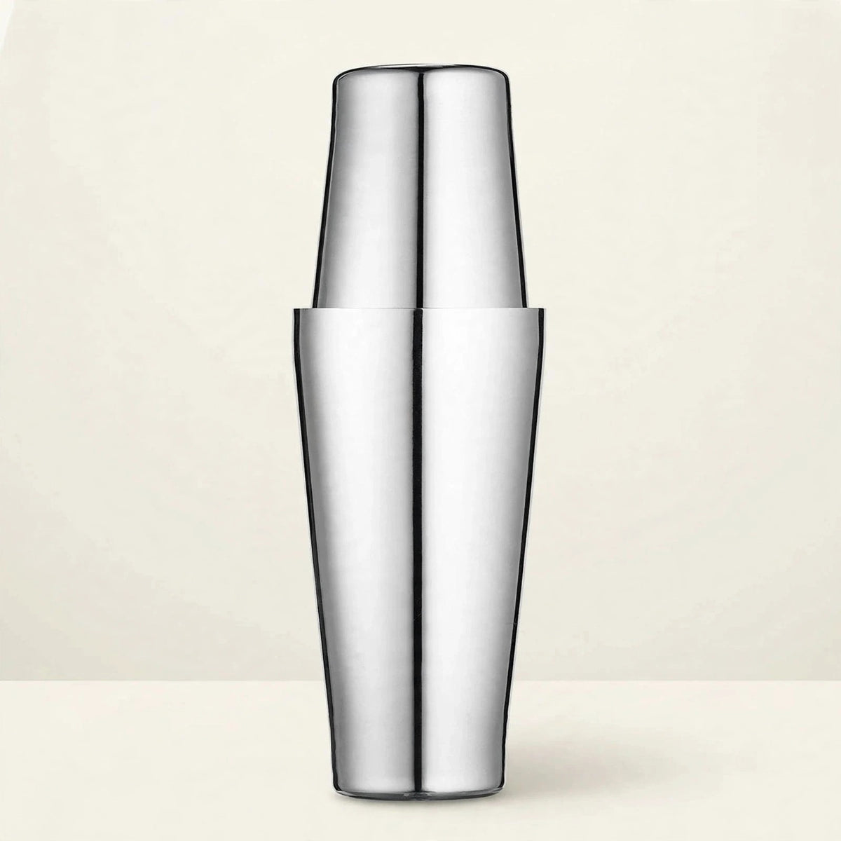 Polished stainless steel cocktail shaker with two tins fitted together stands upright on a cream surface against a pale cream background.