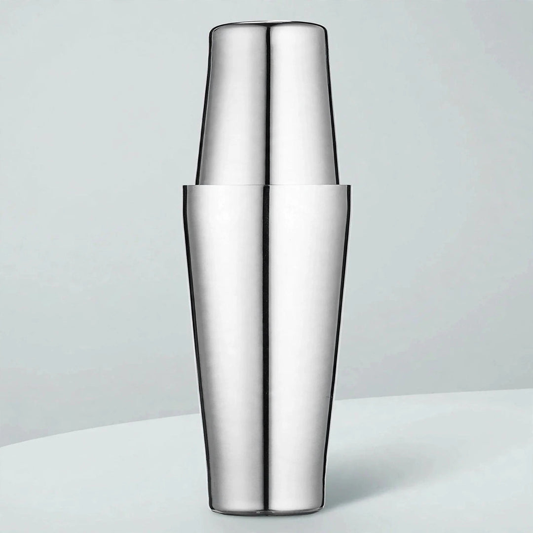 Polished stainless steel cocktail shaker with two tins fitted together stands upright on a pale surface against a light blue-gray background.