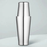 Polished stainless steel cocktail shaker with two tins fitted together stands upright on a pale surface against a light blue-gray background.