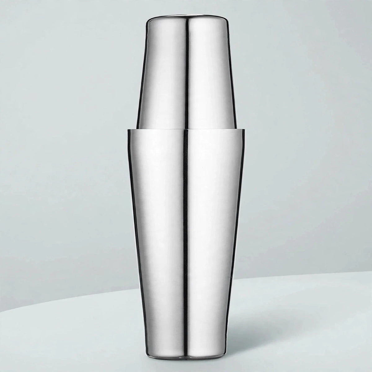 Polished stainless steel cocktail shaker with two tins fitted together stands upright on a pale surface against a light blue-gray background.