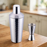 Hammered silver stainless steel cocktail shaker and matching double jigger standing on a wooden kitchen countertop.