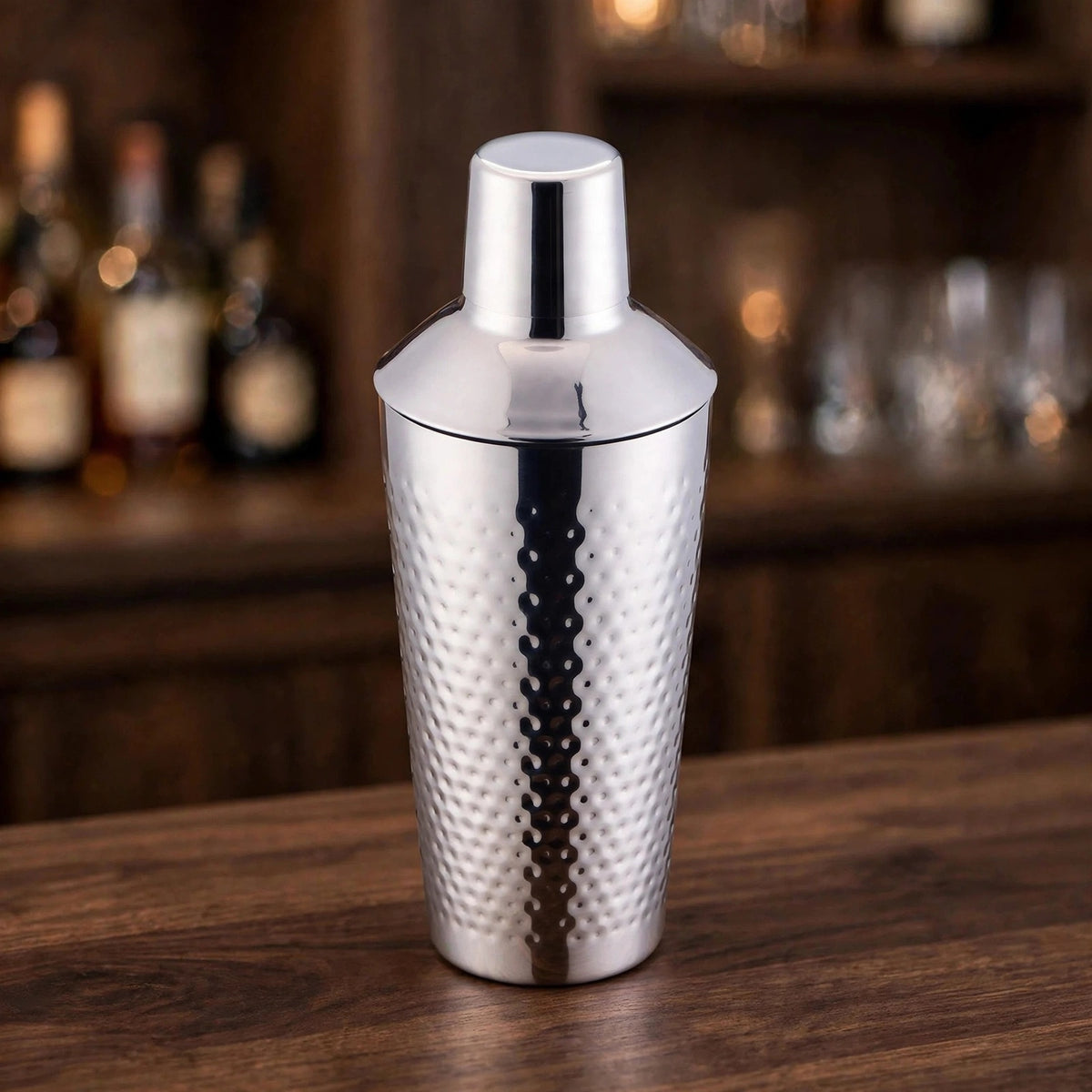 A stainless steel cocktail shaker with a hammered texture, standing on a wooden bar counter with a blurred bar background.