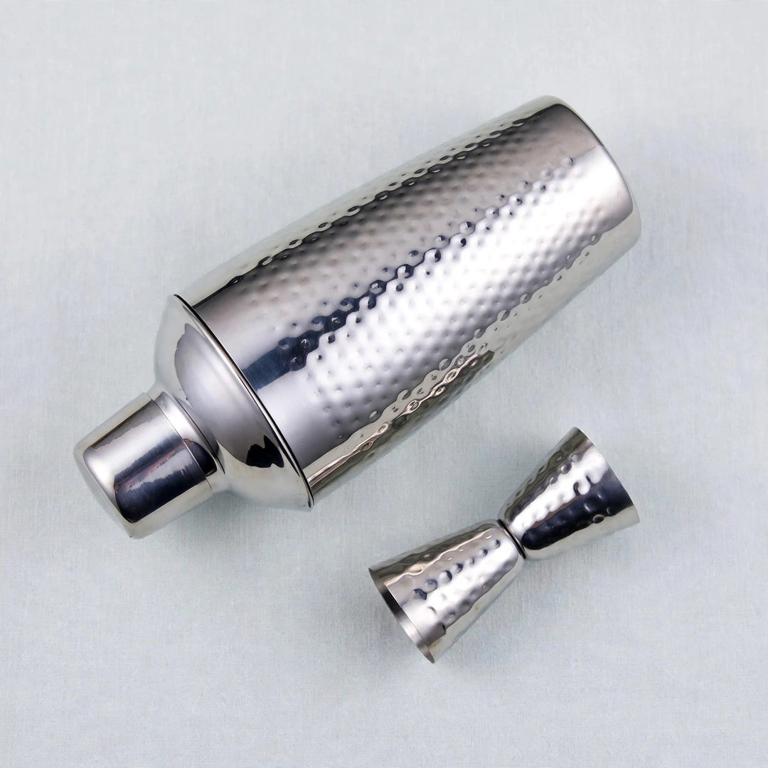 A silver hammered metal cocktail shaker with its matching jigger accessory, displayed on a light gray textured background.