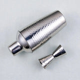 A silver hammered metal cocktail shaker with its matching jigger accessory, displayed on a light gray textured background.
