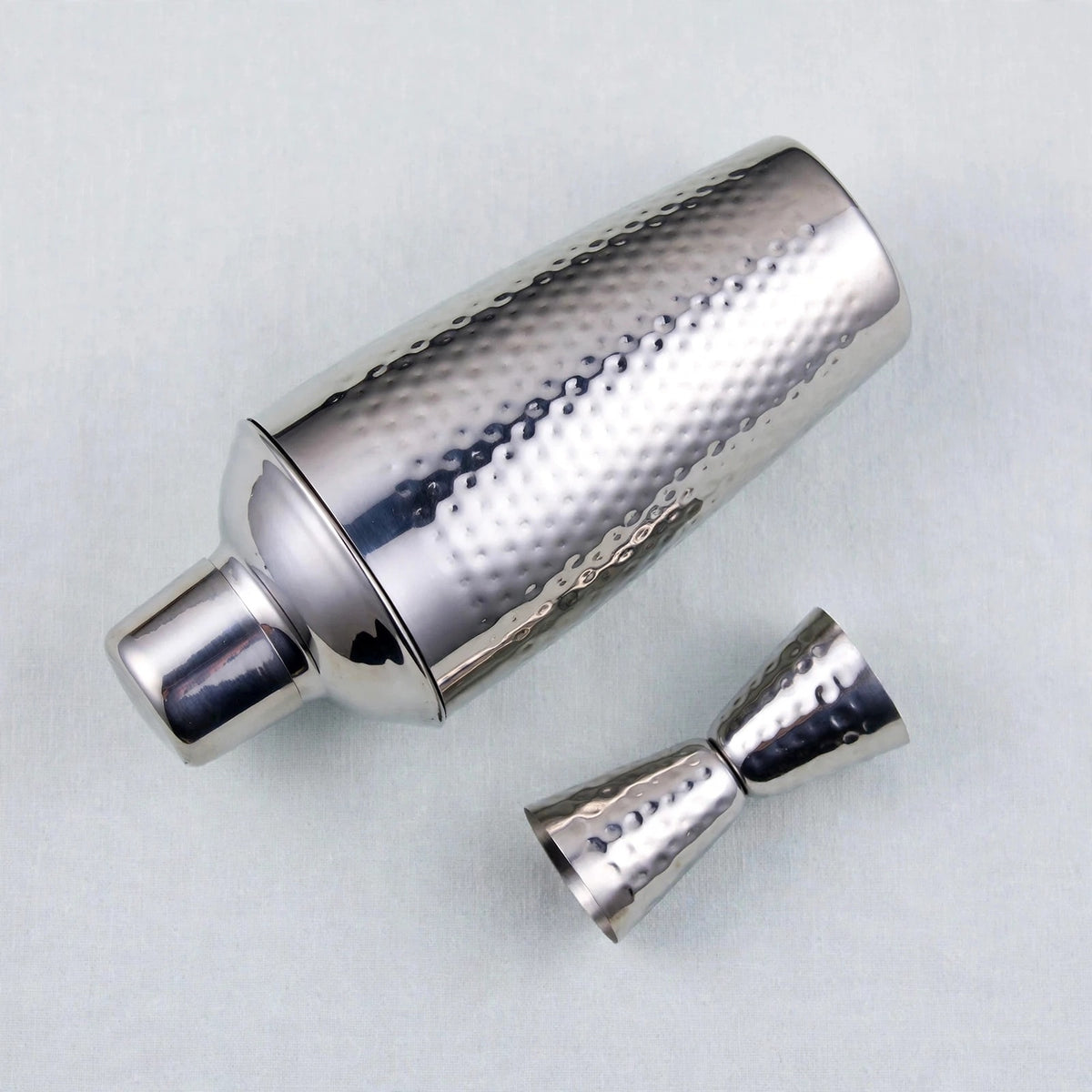 A silver hammered metal cocktail shaker with its matching jigger accessory, displayed on a light gray textured background.