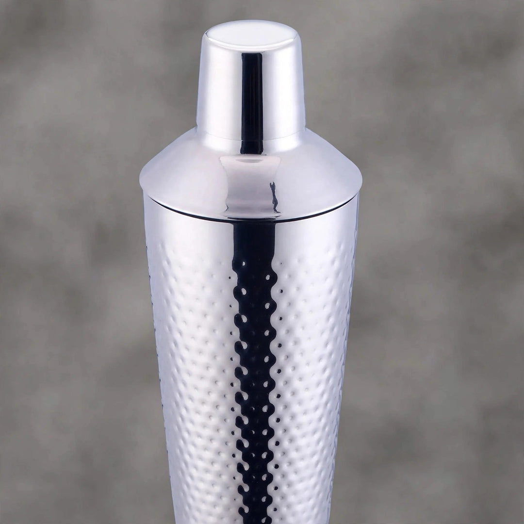 A polished silver stainless steel cocktail shaker with a textured hammered pattern on the body and a matching lid, shown against a soft gray background.