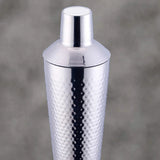 A polished silver stainless steel cocktail shaker with a textured hammered pattern on the body and a matching lid, shown against a soft gray background.