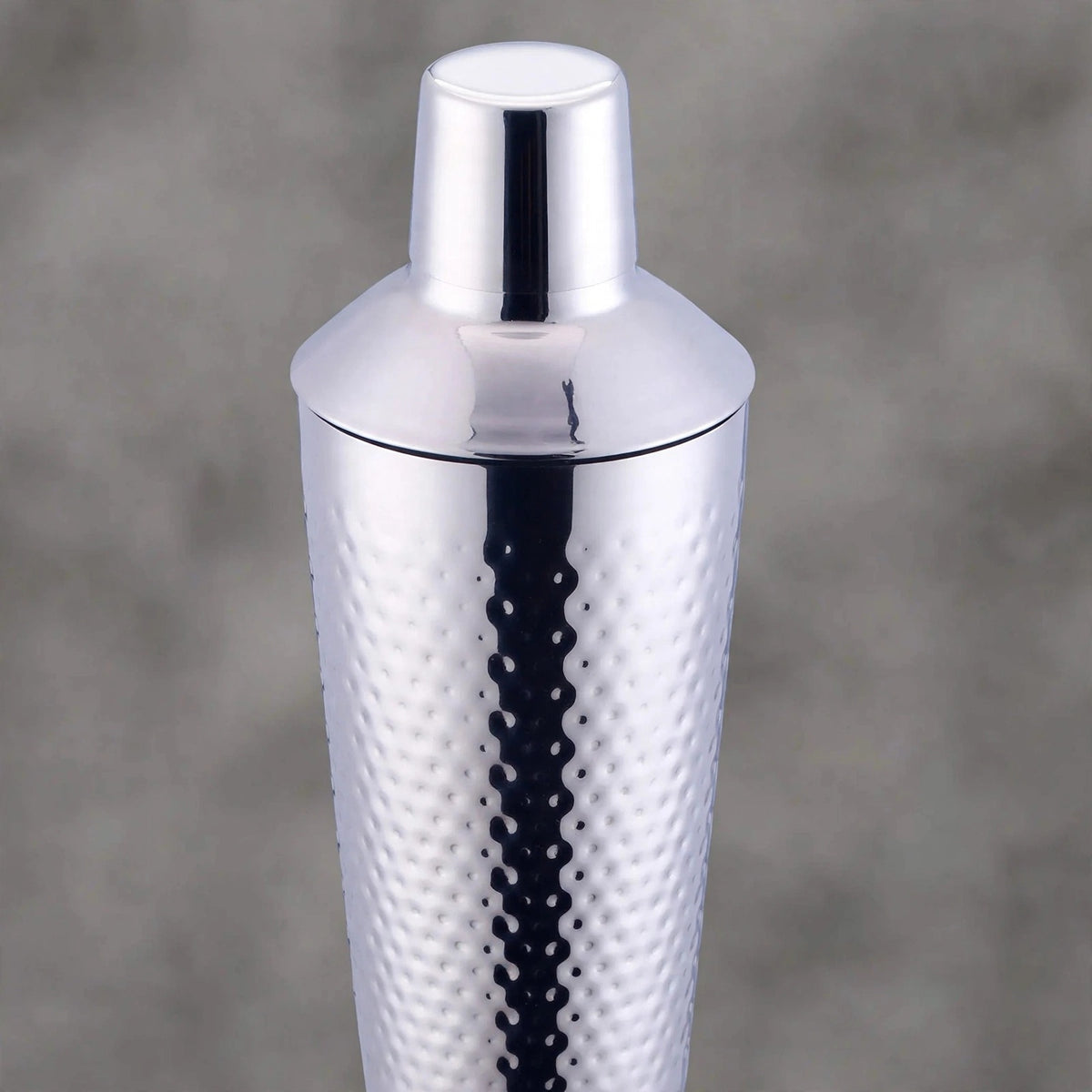 A polished silver stainless steel cocktail shaker with a textured hammered pattern on the body and a matching lid, shown against a soft gray background.