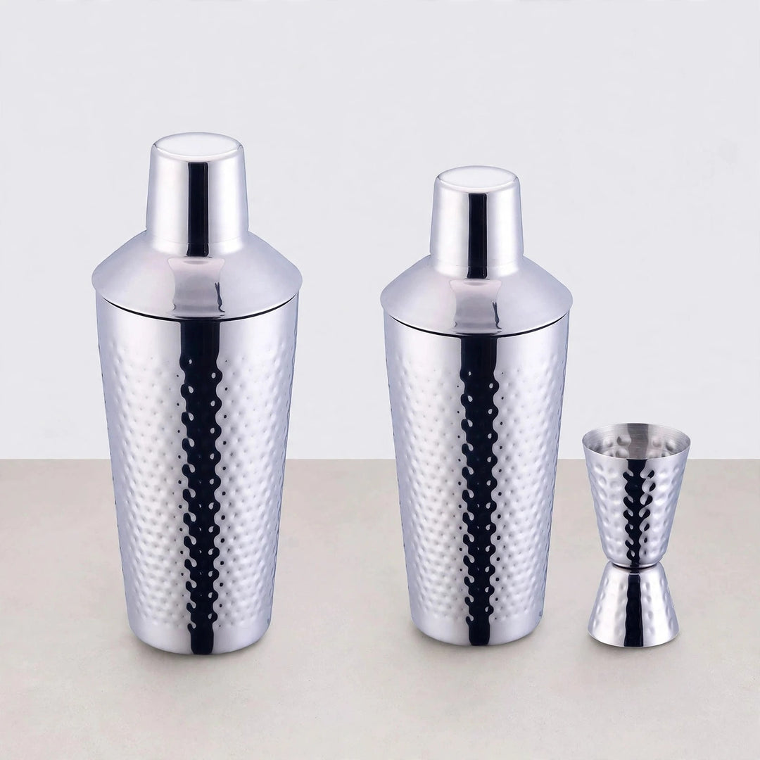 Two hammered silver cocktail shakers in different sizes beside a matching double jigger on a light background.