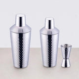 Two hammered silver cocktail shakers in different sizes beside a matching double jigger on a light background.