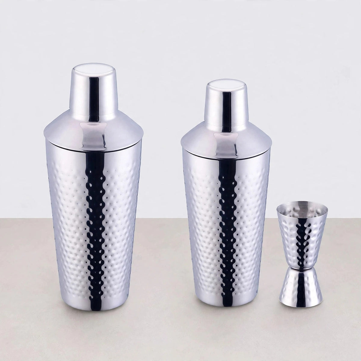 Two hammered silver cocktail shakers in different sizes beside a matching double jigger on a light background.