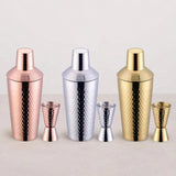 Three hammered cocktail shaker and jigger sets in copper, silver, and gold lined up on a white surface.