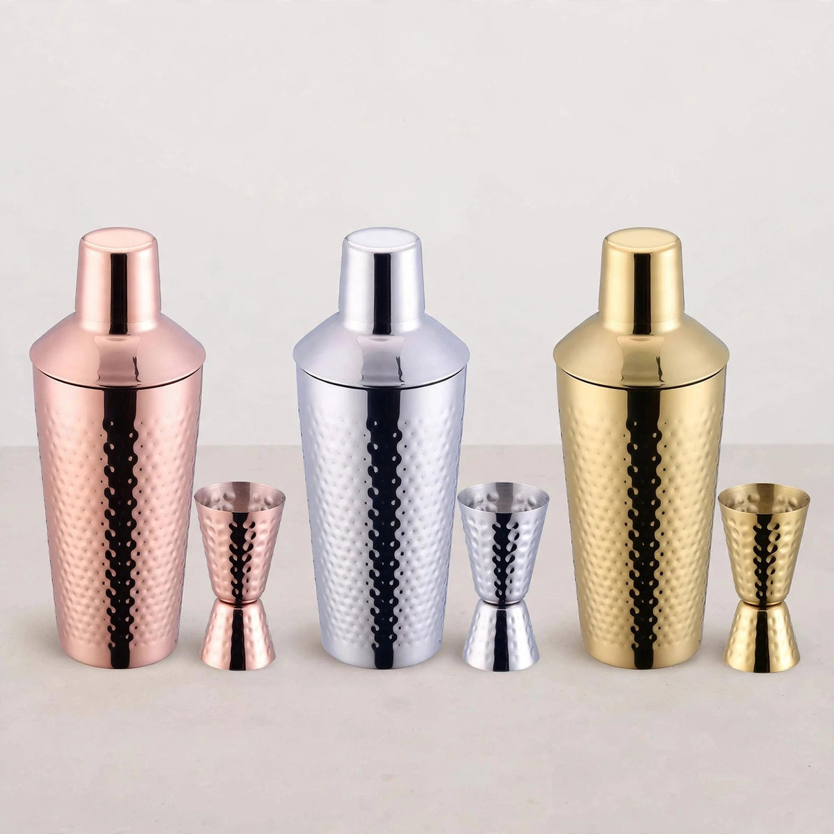 Three hammered cocktail shaker and jigger sets in copper, silver, and gold lined up on a white surface.