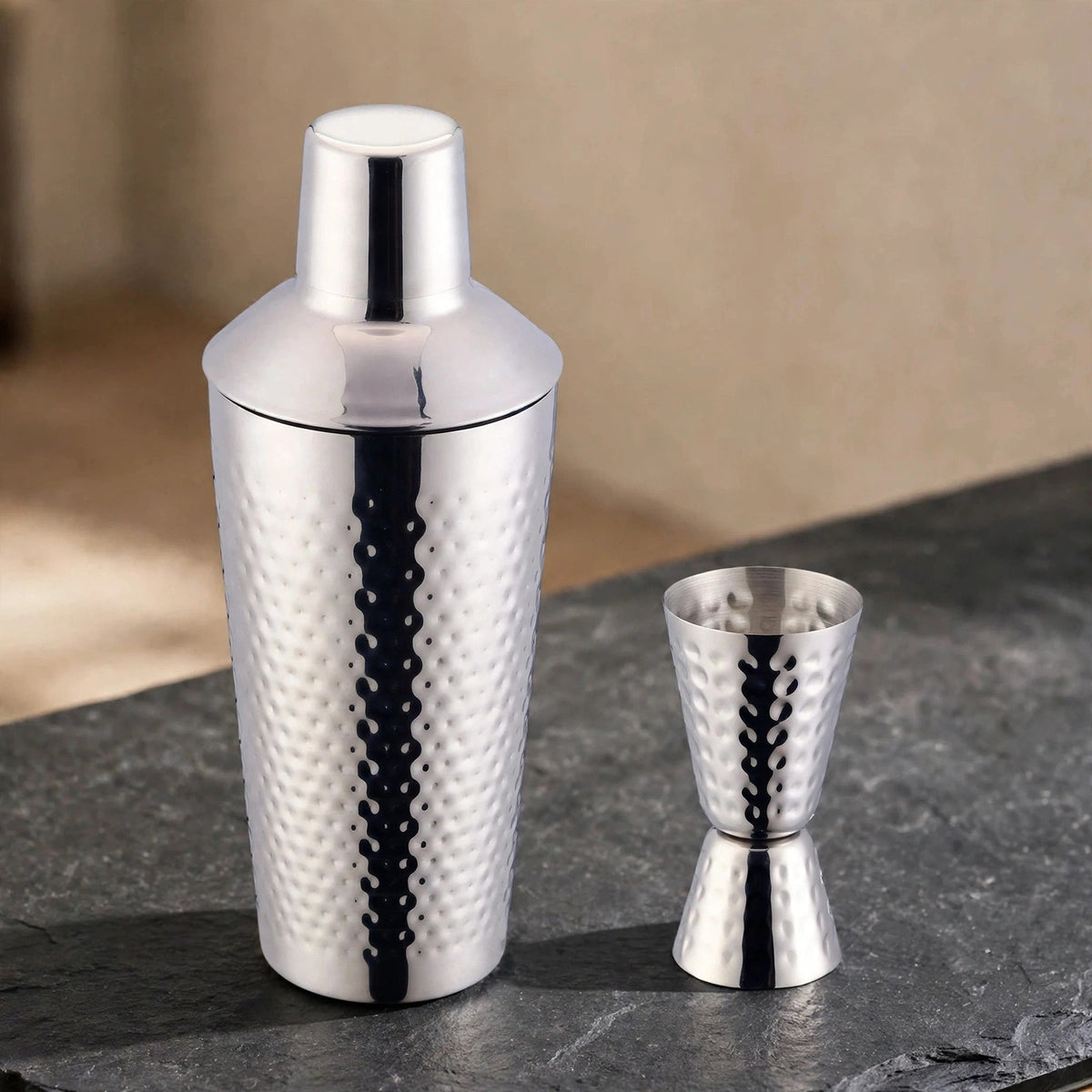 Hammered silver cocktail shaker and double jigger placed on a dark slate stone serving surface.