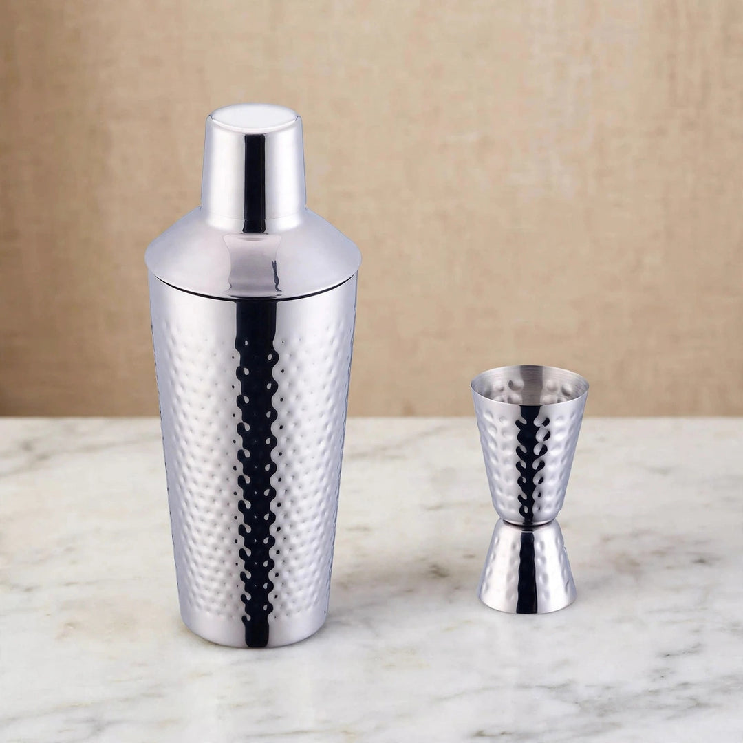 Silver hammered cocktail shaker and double jigger set on a white marble surface against a beige wall.