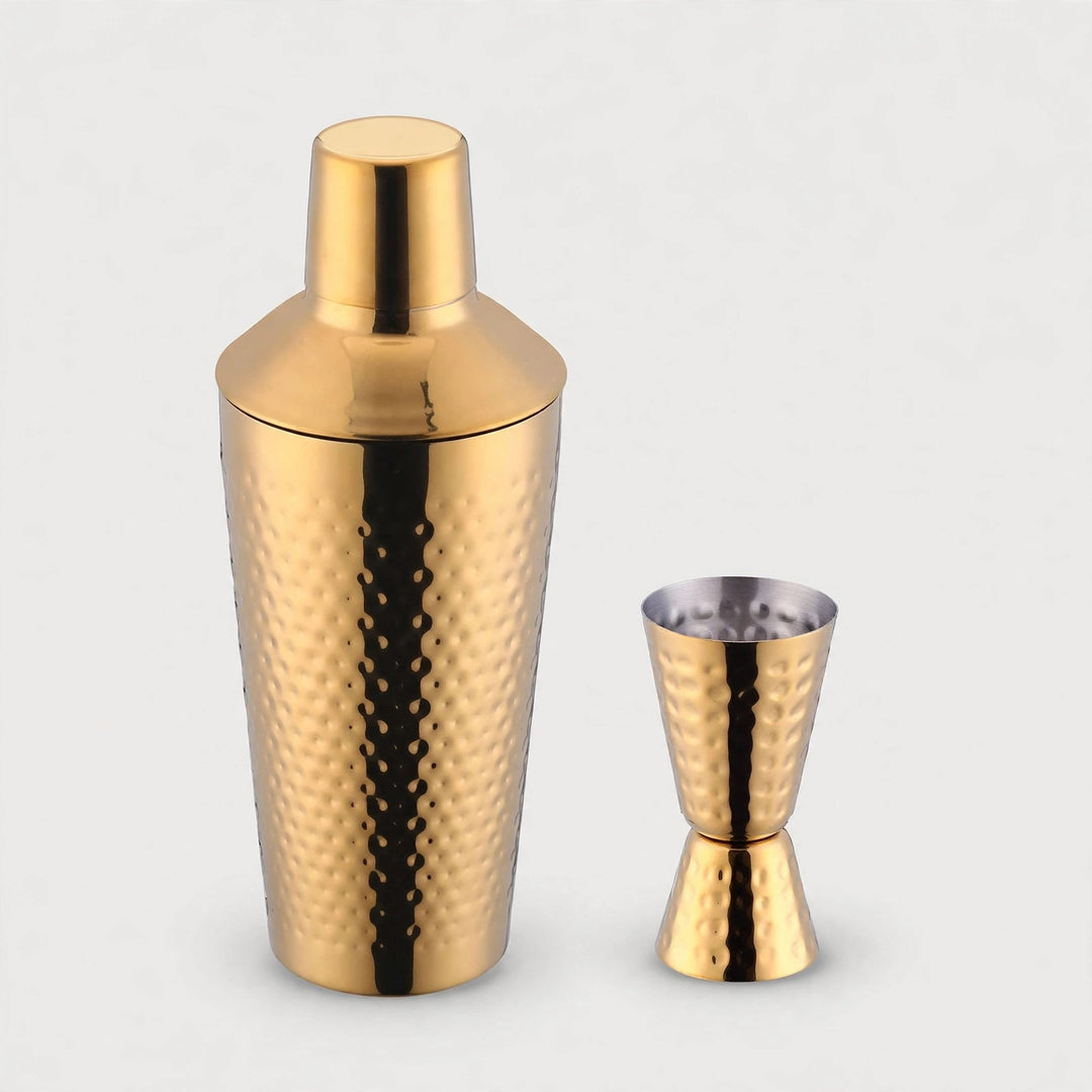 Gold hammered metal cocktail shaker with a smooth lid stands beside a matching double-ended jigger against a light grey background.