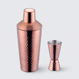 Rose gold hammered metal cocktail shaker with a smooth lid stands beside a matching double-ended jigger against a light grey background.