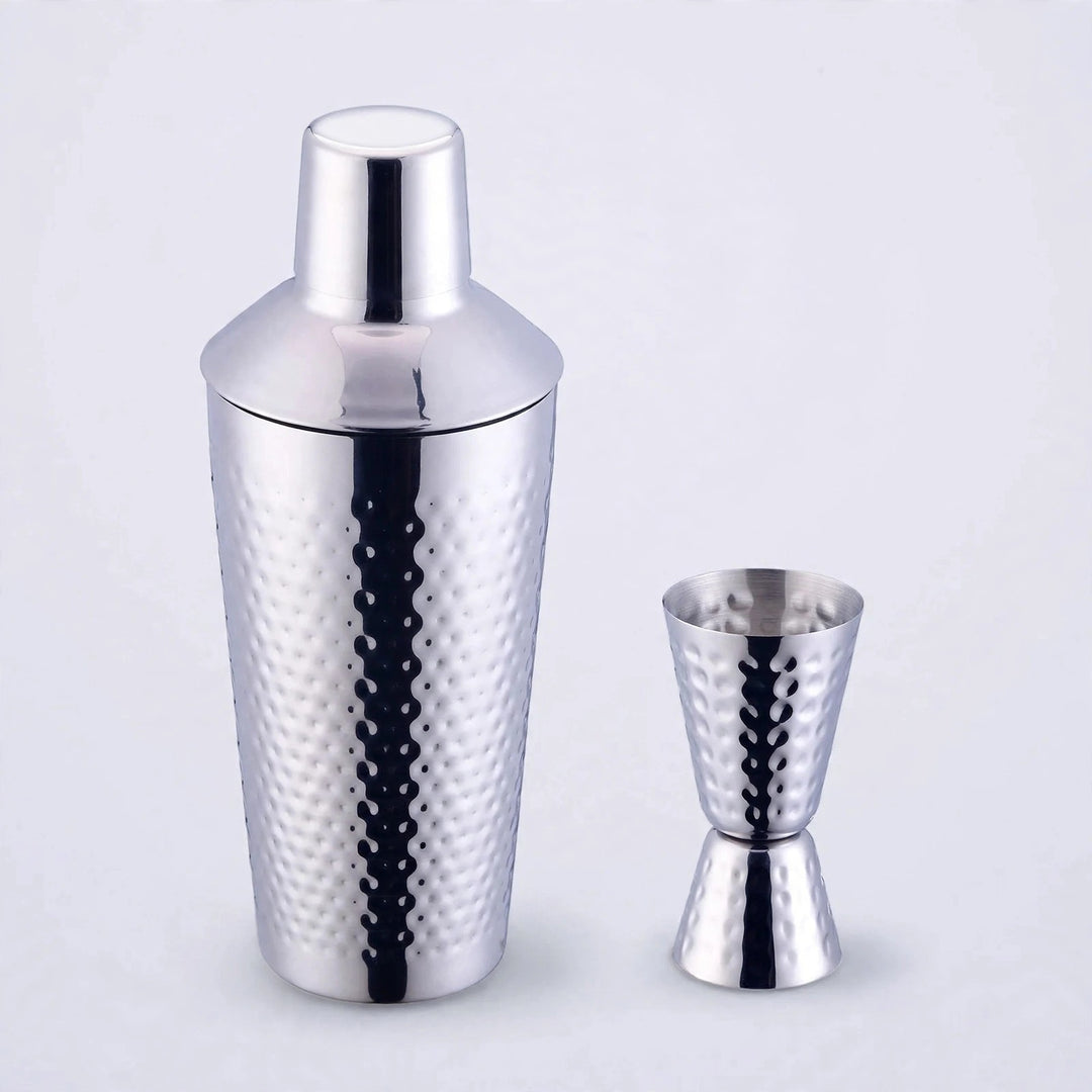 Silver hammered stainless steel cocktail shaker with matching double jigger on a plain light studio background.