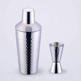 Silver hammered stainless steel cocktail shaker with matching double jigger on a plain light studio background.